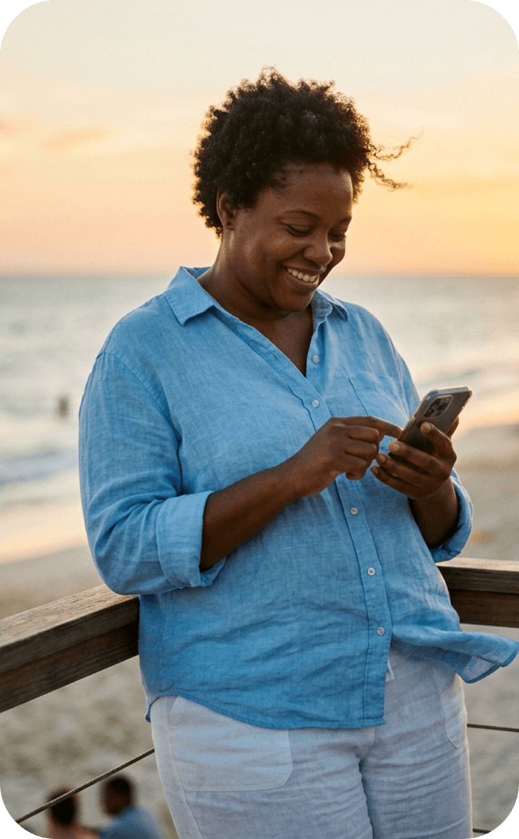 A woman smiling at her phone on a boardwalk by the beach at sunset